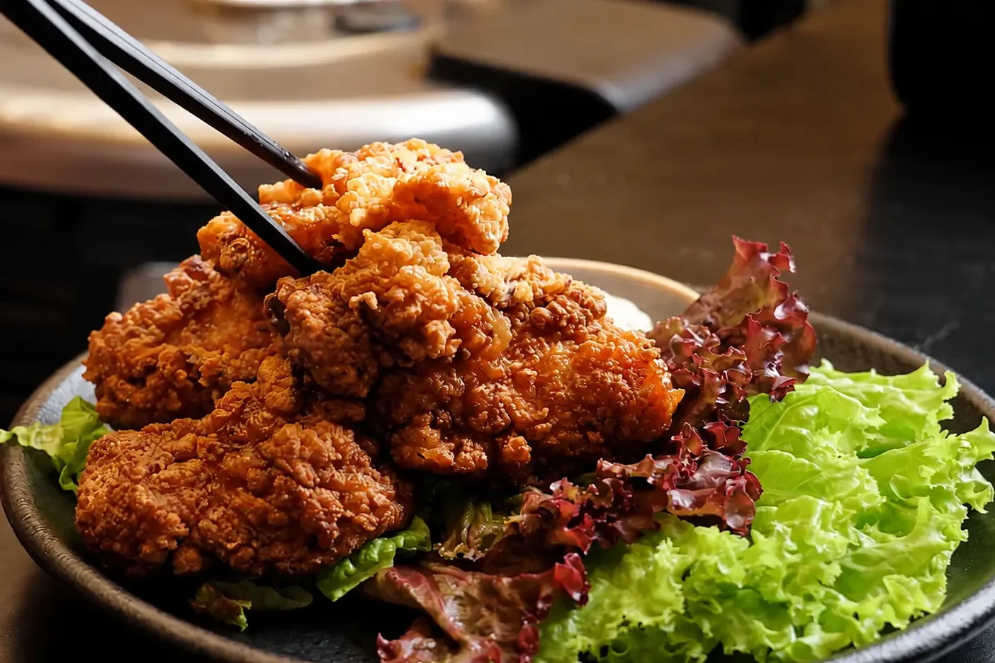 A plate of crispy fried chicken served with fresh green and red lettuce, with chopsticks lifting a piece of the dish.